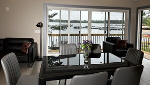 A modern living room with a glass table, leather chairs, a black sofa, and a view of a lake through large windows.