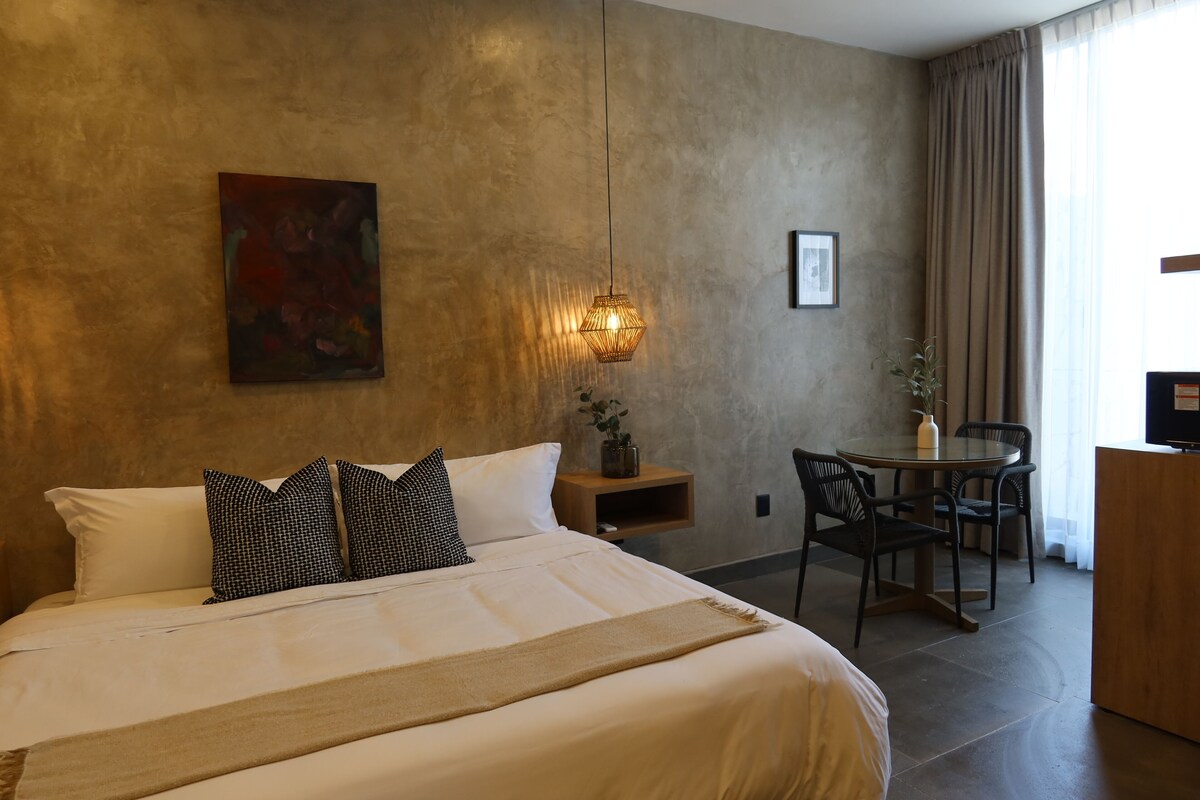 Quarto de hotel com uma cama grande, uma escrivaninha com cadeira, uma luminária de parede e um quadro.