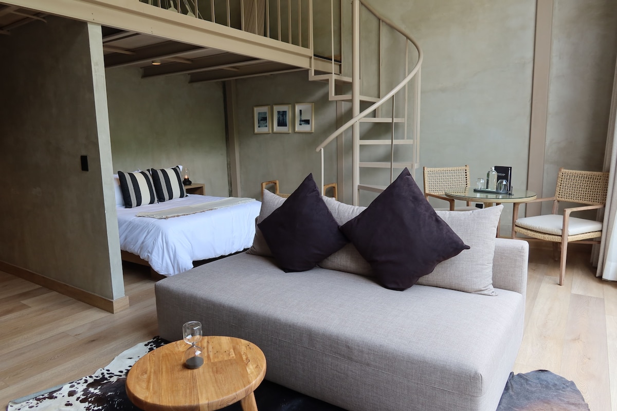 Quarto moderno com sofá, mesinha redonda de vidro, cama com edredom branco e escada.