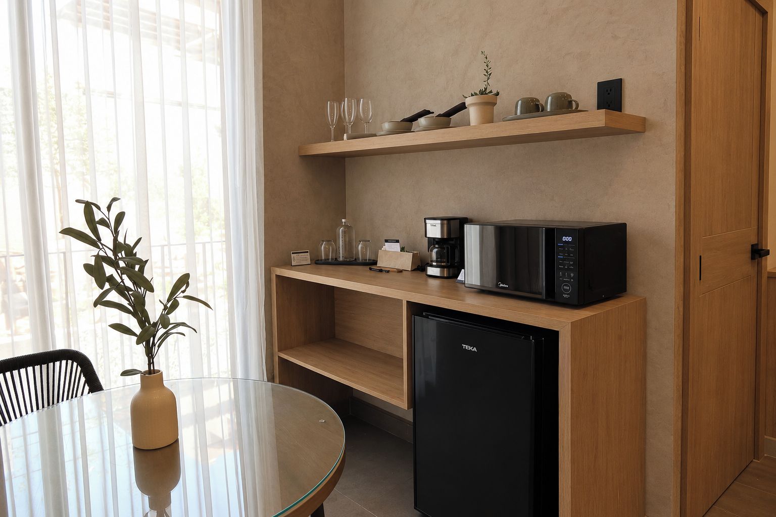 Uma kitchenette compacta com micro-ondas, máquina de café e pequena geladeira. Há uma mesa de vidro com um vaso de plantas verdes e uma cadeira.
