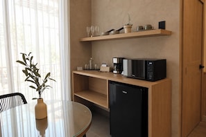 Cocina compacta equipada con microondas, cafetera y pequeño refrigerador. Hay una mesa de vidrio con un florero de plantas verdes y una silla.