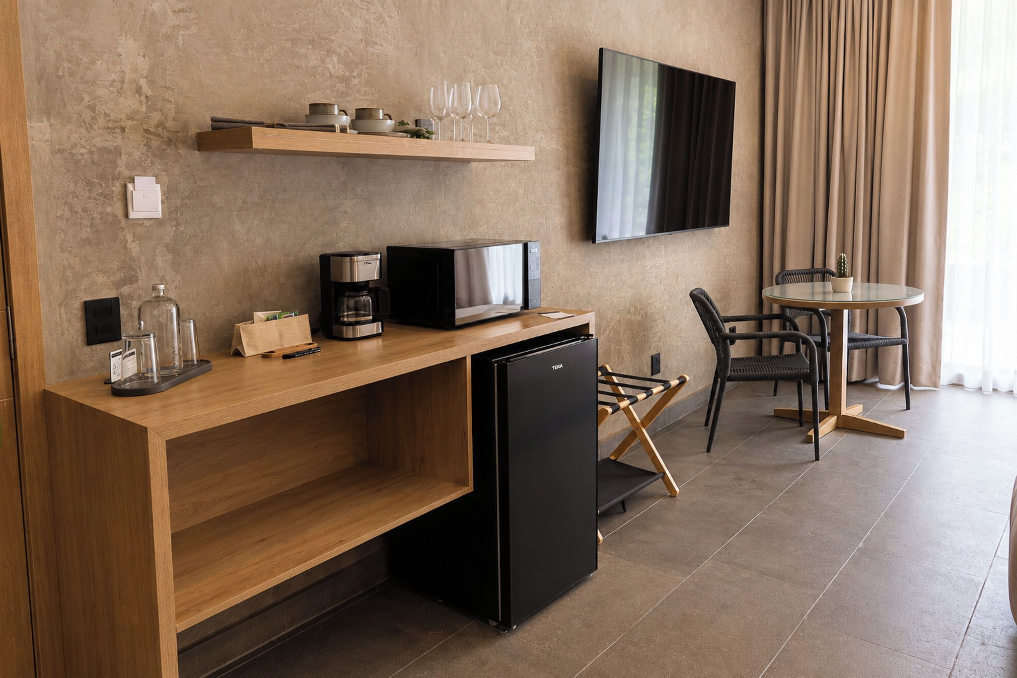 A modern kitchenette with a coffee maker, microwave, and refrigerator. There is a wooden shelf above the counter holding cups and glasses. A flat-screen TV is mounted on the wall, and a dining area with chairs and a table is visible.