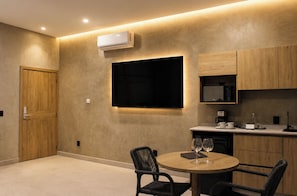 A modern living room with a flat-screen TV, a round dining table with chairs, a kitchen area with cabinets, and an air conditioning unit.