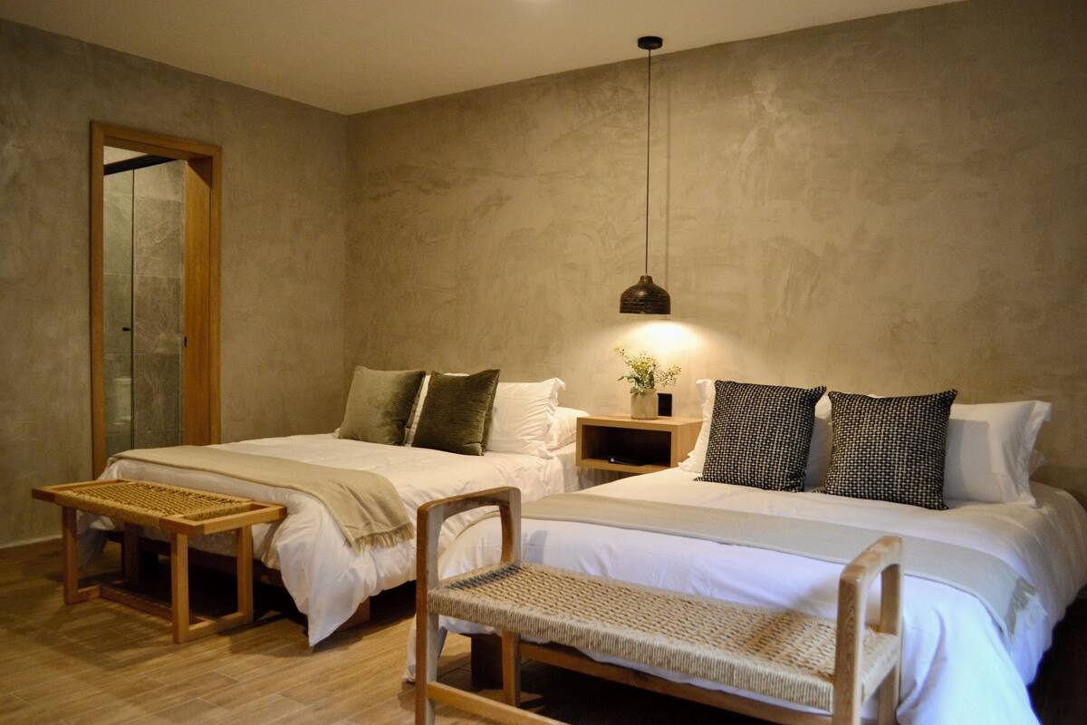 Quarto com duas camas, um banco de madeira, mesa de cabeceira com abajur e espelho na parede.