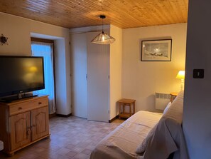A room with a television on a wooden cabinet, a bed with a white duvet, a small wooden table, a hanging light fixture, and a framed picture on the wall.