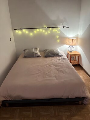 A bedroom with a bed, pillows, a bedside table with a lamp, and string lights on the wall.