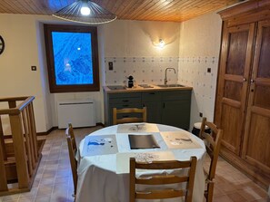 A dining area with a table set for two, a kitchen with a sink and cabinets, and a large window with a view of a blue sky.