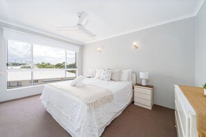 A bedroom with a bed, a nightstand, a lamp, a ceiling fan, and a window with a view. - 3BR Apt + Pool Great Location Opp Beach & Park (Urangan)