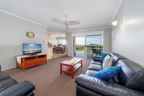 A living room with a TV, a wooden coffee table, a black leather sofa, and a balcony with a view. - 3BR Apt + Pool Great Location Opp Beach & Park (Urangan)