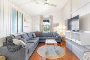 Ein Wohnzimmer mit einem grauen Ecksofa, einem kleinen Holztisch, einem Fernseher auf einem weißen Schrank und einem Deckenventilator.