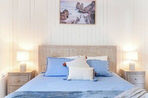 Ein Bett mit Holz-Kopfteil, zwei Nachttische mit Lampen und ein gerahmtes Bild einer Küstenlandschaft an der Wand.