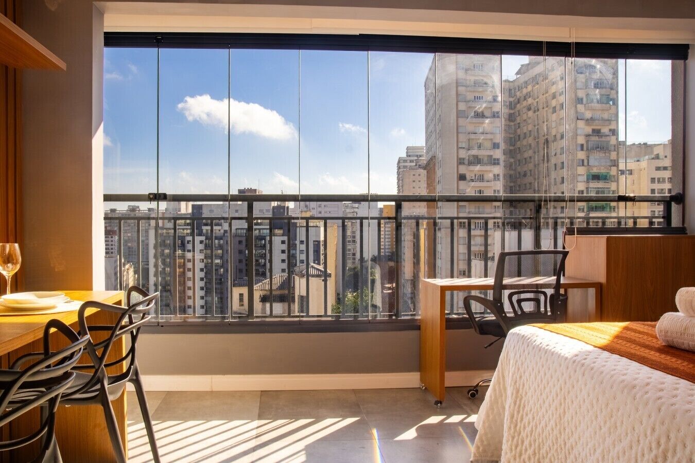 A hotel room with a large window, a dining table with chairs, a bed, and a view of high-rise buildings.