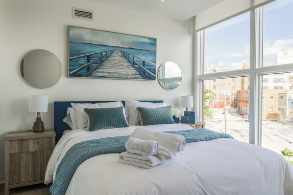 A hotel room with a large bed, a painting of a pier, a view of buildings, and a window. - Luxury High Rise Walkable Free Parking + Views (Kansas City)