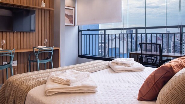 Ein Hotelzimmer mit Bett, zwei Stühlen, einem kleinen Tisch, einem Fernseher und einem Balkon mit Blick auf die Stadt.