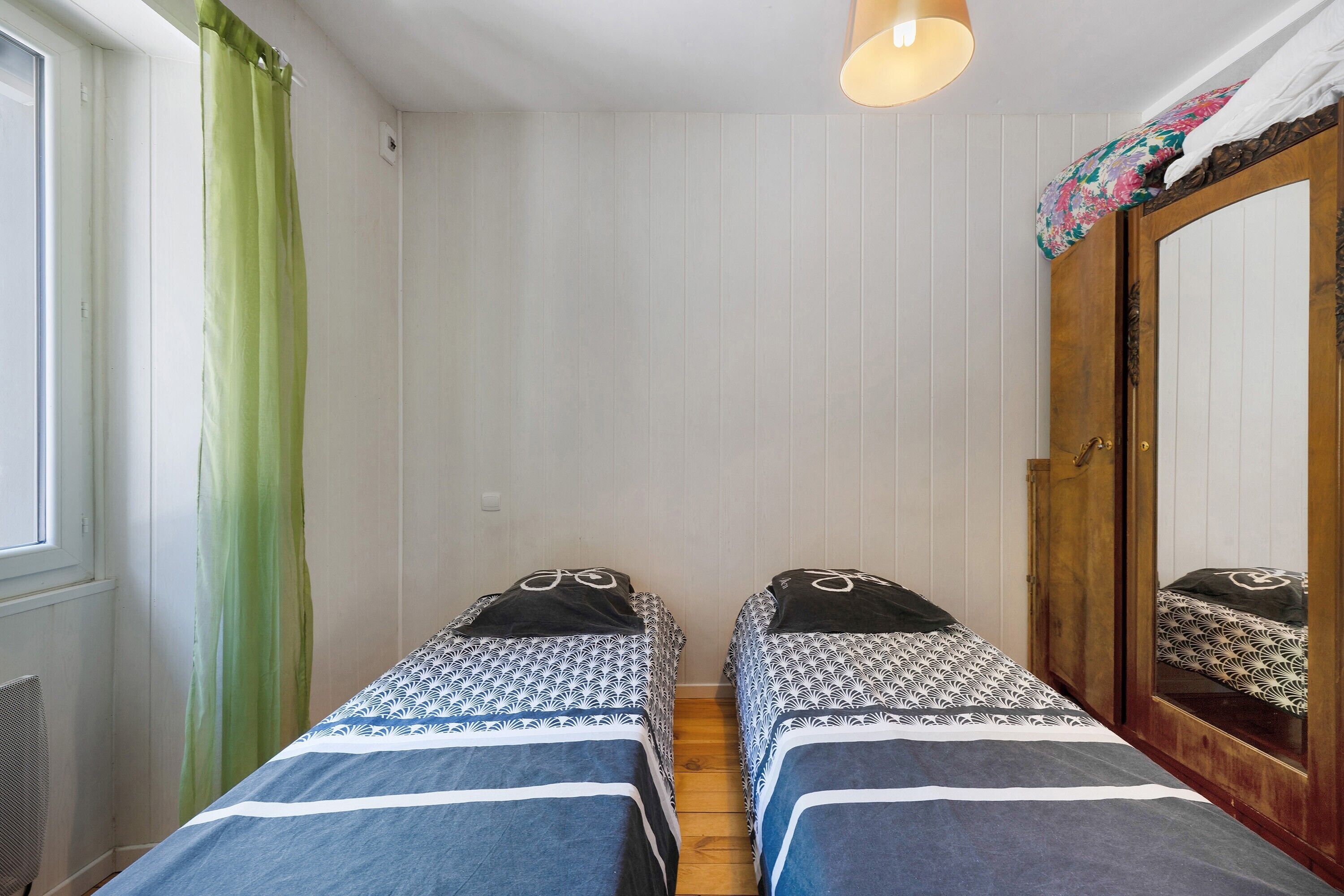 Une chambre avec deux lits simples, une armoire en bois, un miroir et une fenêtre avec un rideau.