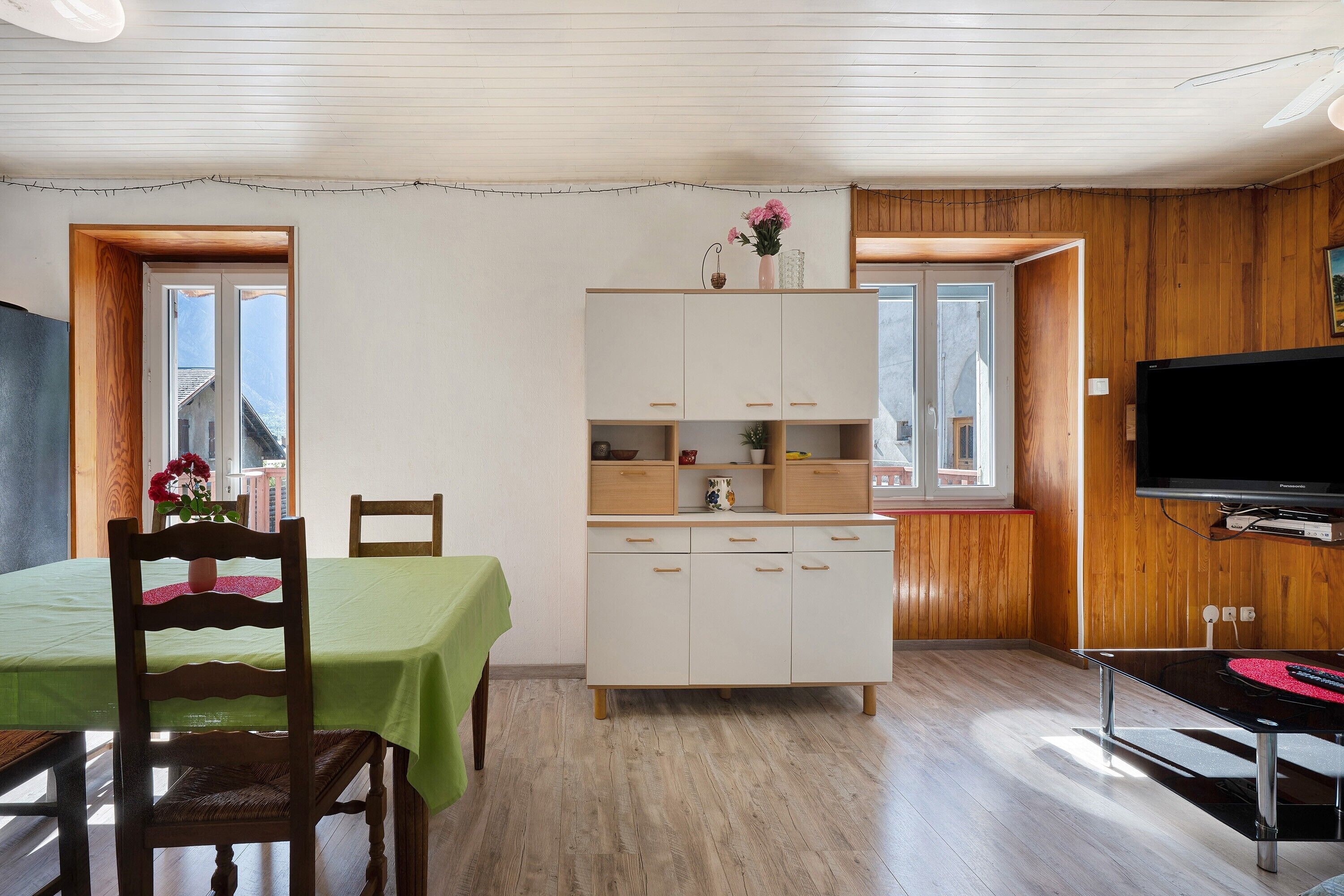 Ein Essbereich mit Tisch und Stühlen, eine weiße Kommode mit Schubladen, ein an der Wand befestigter Fernseher und ein Fenster mit Blick auf Gebäude.