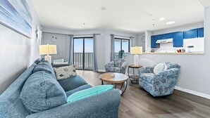 Una sala de estar con un sofá, dos sillones, una mesa de centro y una zona de cocina con armarios azules.