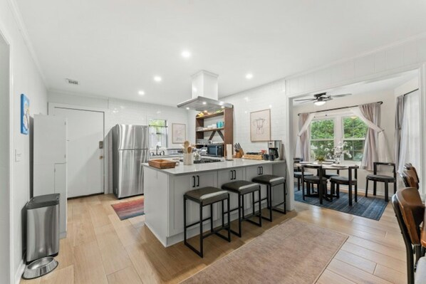 A modern kitchen with a central island, bar stools, and a dining area with a view of the outdoors. - Robin's Nest - Private Pool Retreat (North Myrtle Beach)