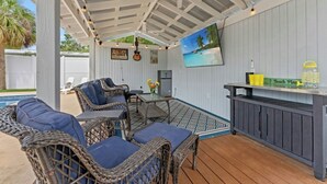 Een overdekt terrasgedeelte met rieten meubels, een tafel en een bargedeelte.