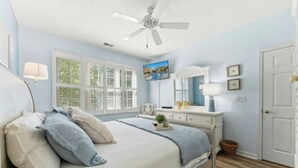 Une chambre à coucher comprenant un lit, un ventilateur de plafond, une télévision, une commode et une fenêtre avec des stores.