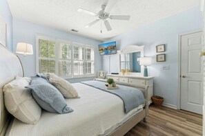 Ein Schlafzimmer mit Bett, Deckenventilator, Fernseher, Schminktisch und Fenster mit Jalousien.