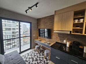 A modern kitchen with a large window, a flat-screen TV, and a dining area with stools. - Studio Clean - Pinheiros (São Paulo)
