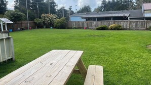Ein Holztisch für ein Picknick in einem Garten mit einem grünen Rasen, einem Holzzaun und einem Gebäude mit einem Metall-Dach im Hintergrund.