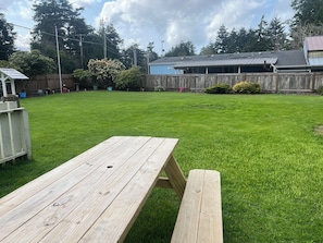 Ein Holztisch für ein Picknick in einem Garten mit einem grünen Rasen, einem Holzzaun und einem Gebäude mit einem Metall-Dach im Hintergrund.