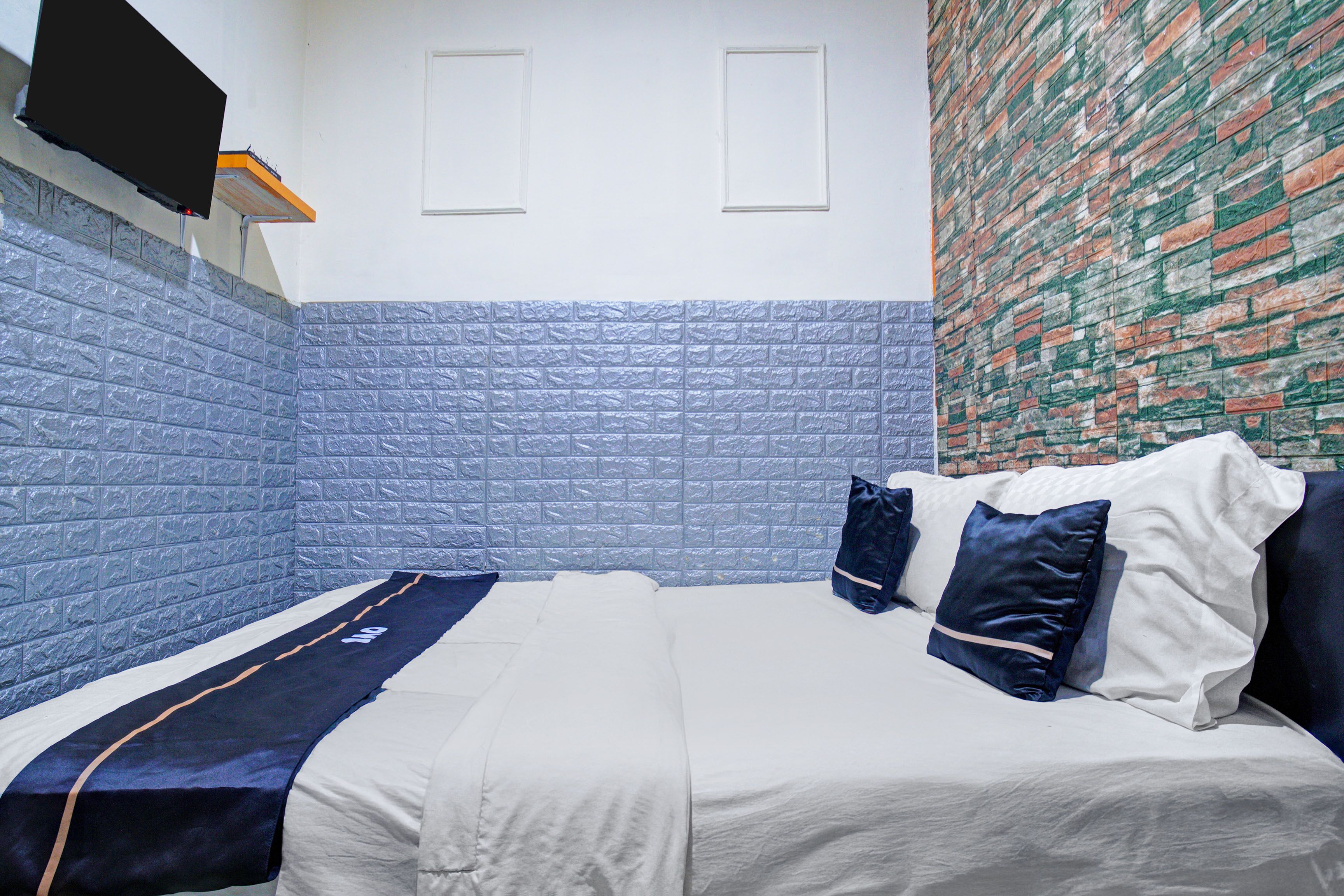 Een slaapkamer met een bed, kussens, een aan de muur bevestigde televisie en een muur met een bakstenen patroon.