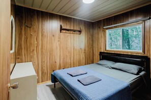 Une chambre avec des murs en bois, un lit recouvert d’une couette bleue, une fenêtre donnant sur une forêt et un porte-manteau.