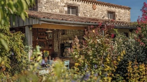 Ein rustikales Steinhaus mit Ziegeldach, einer Holzveranda und einem Garten mit verschiedenen Pflanzen und Blumen.
