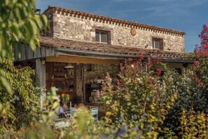Un bâtiment rustique en pierre, doté d’un toit en tuiles, d’un porche en bois et d’un jardin où poussent diverses plantes et fleurs.