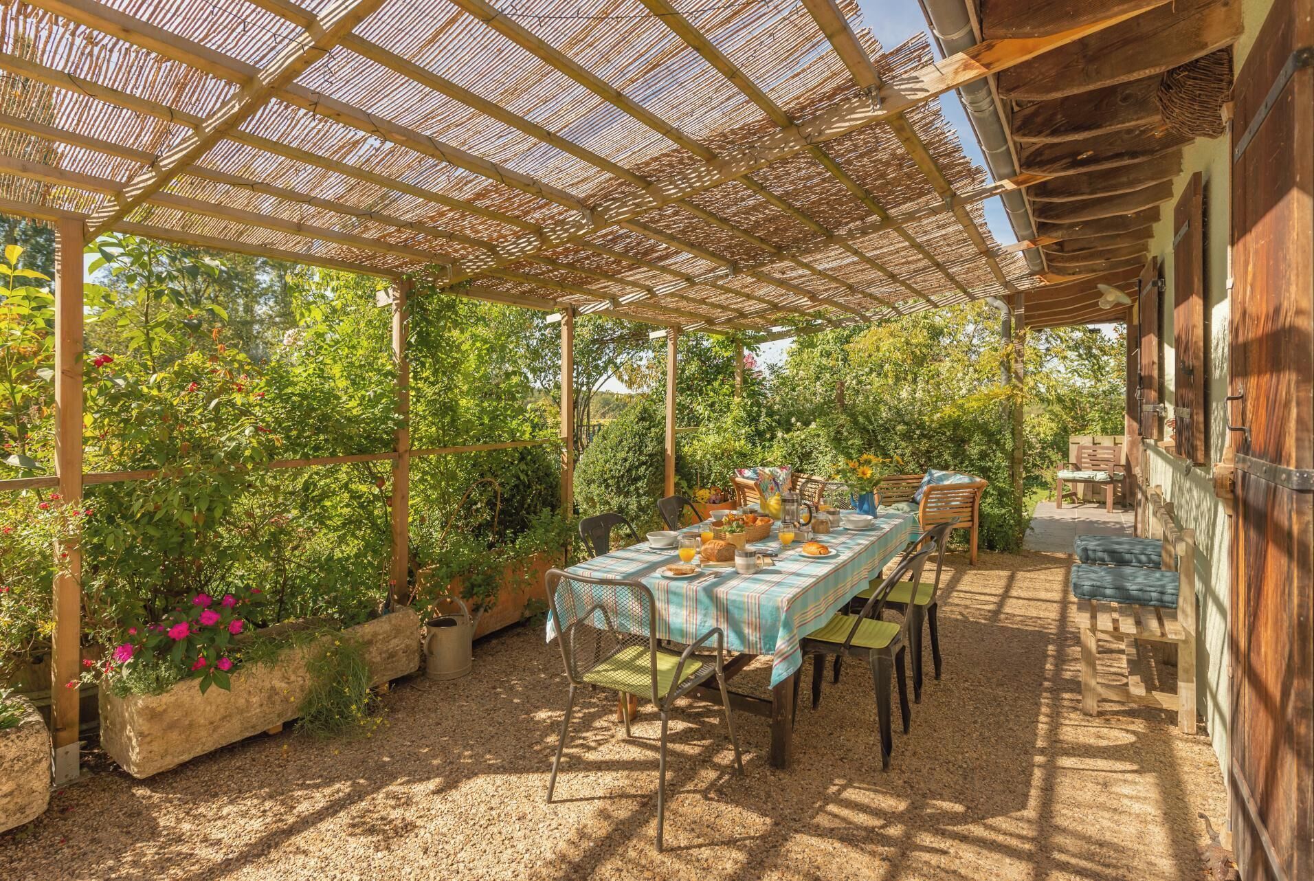 An outdoor patio with a table set for dining, surrounded by greenery and a pergola structure.