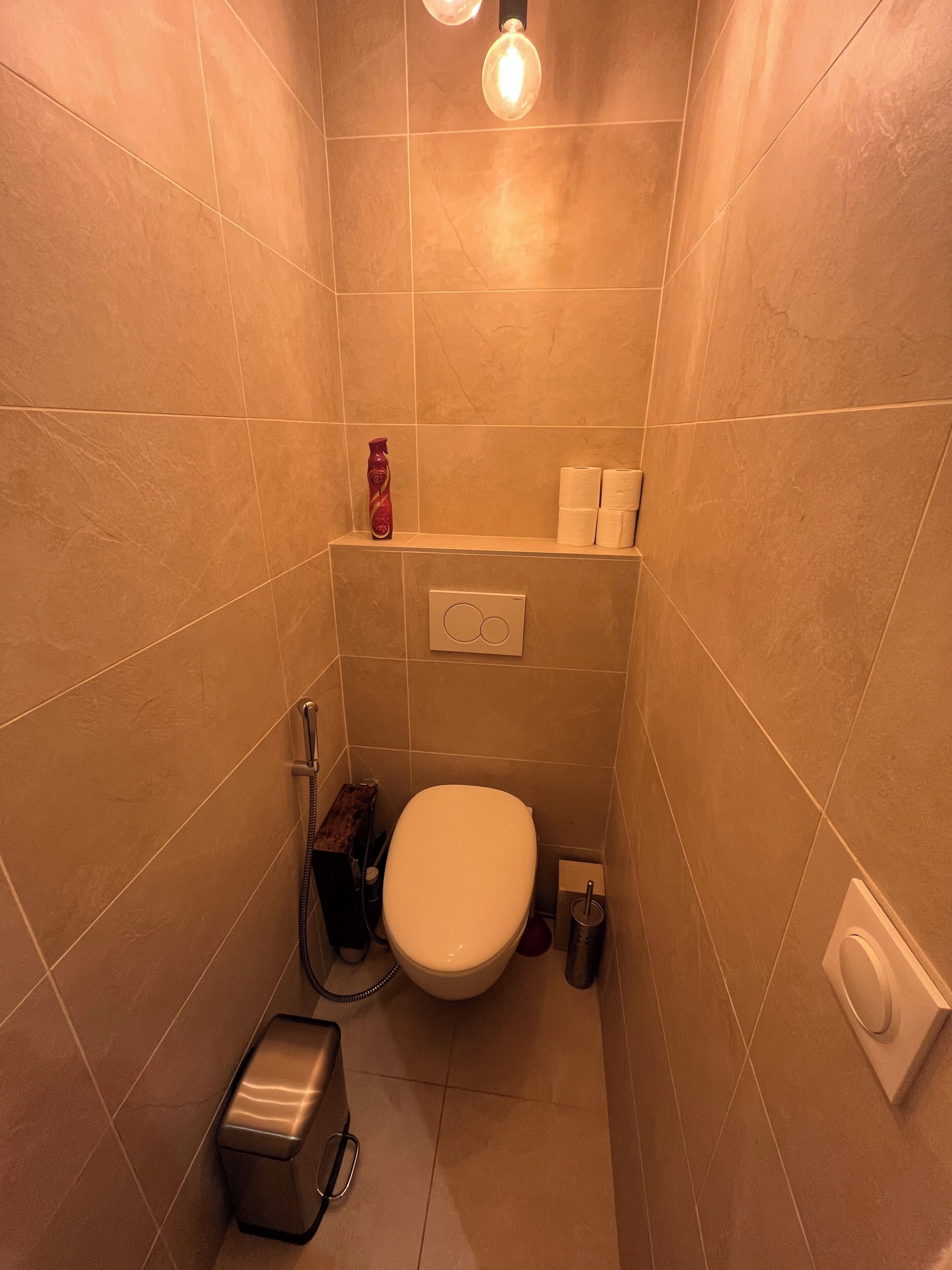 A small, modern bathroom with a toilet, a shelf with toiletries, a towel dispenser, and a hand dryer.