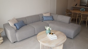 Un salon moderne avec un canapé d’angle gris, une table basse ronde en bois et un coin repas comprenant une table et des chaises en bois.
