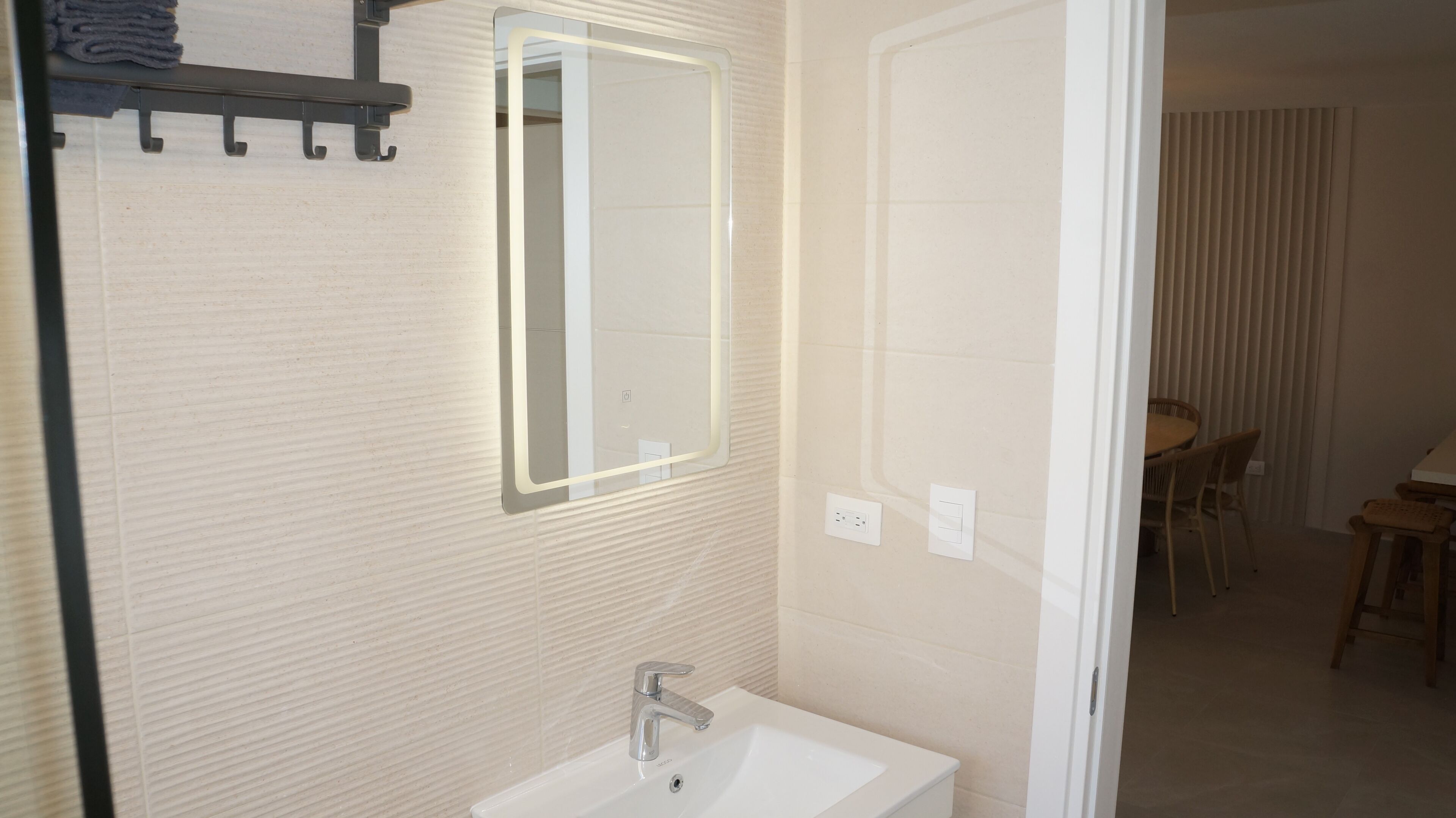 Baño con lavamanos, espejo, toallero y puerta que da a un comedor.