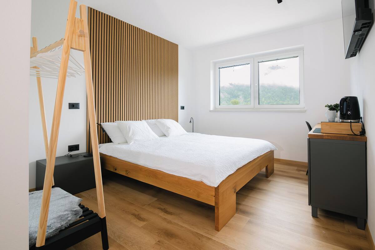 A modern bedroom with a wooden bed, a wooden wardrobe, a small desk, and a window with a view of greenery.