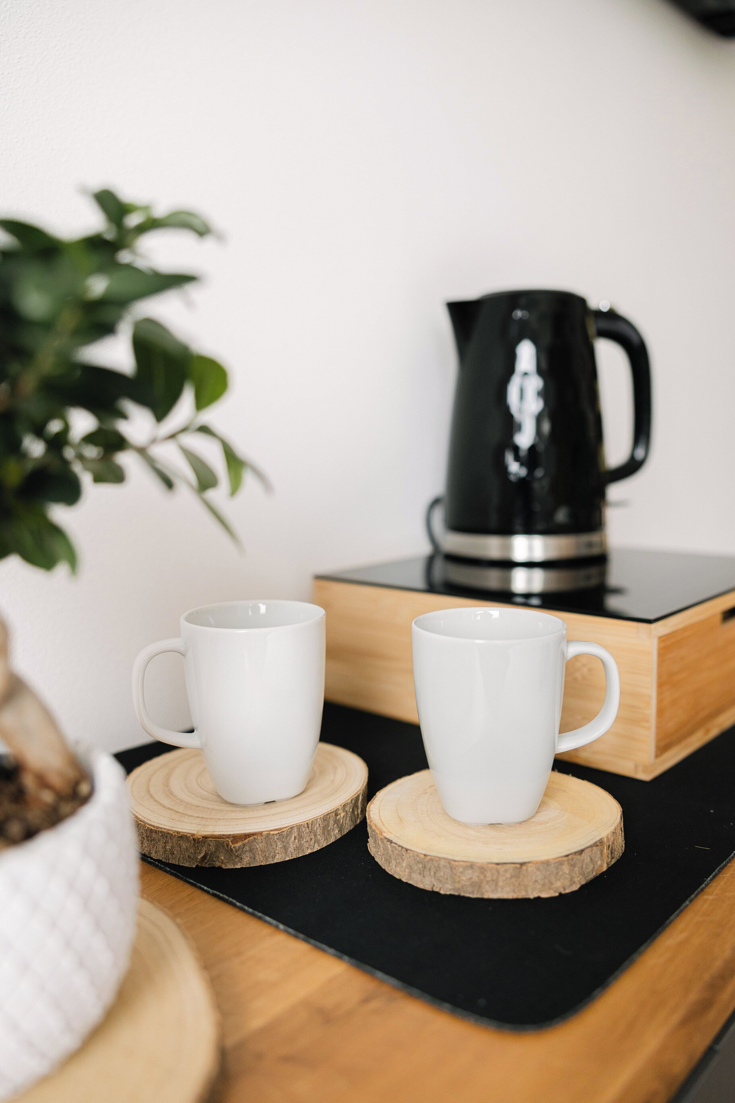 Zwei weiße Tassen auf Holzuntersetzern, eine schwarze Kaffeemaschine und eine Pflanze im Hintergrund.