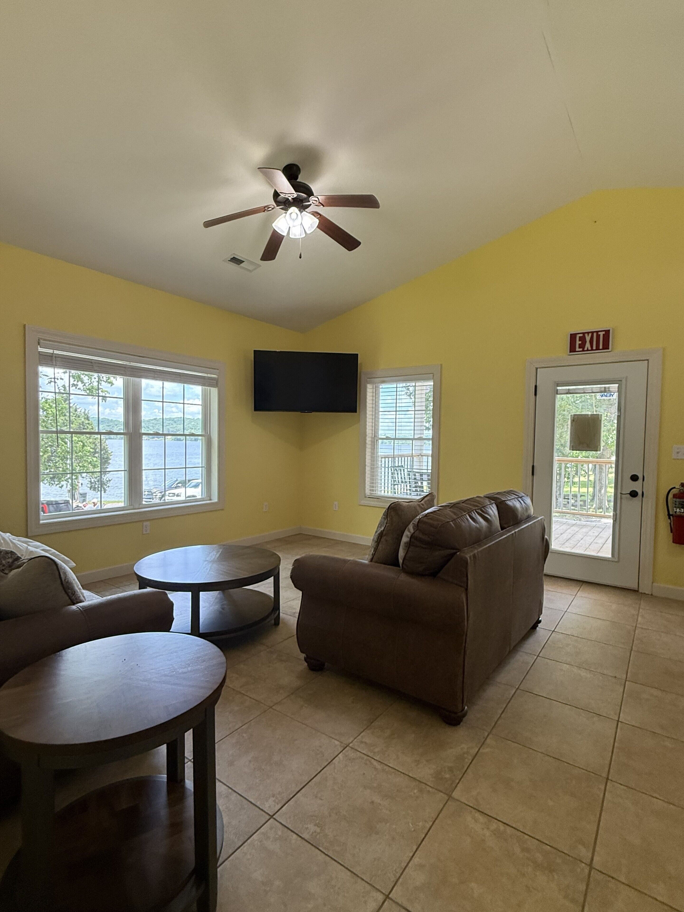 A room with a ceiling fan, a television mounted on the wall, a brown sofa, two round tables, and a door marked 'EXIT'.