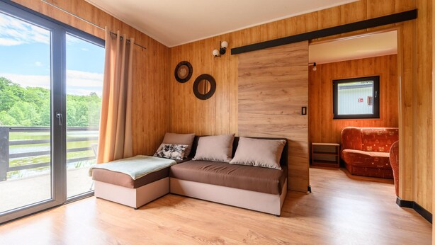 A modern living room with a sectional sofa, a wooden floor, and a large window offering a view of greenery. - Single-story Holiday Home by the Pond no. 3 (Darlowo)