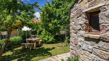 Un bâtiment en pierre avec une fenêtre, un chemin en pierre et une table en bois entourée de chaises, le tout situé dans un jardin.
