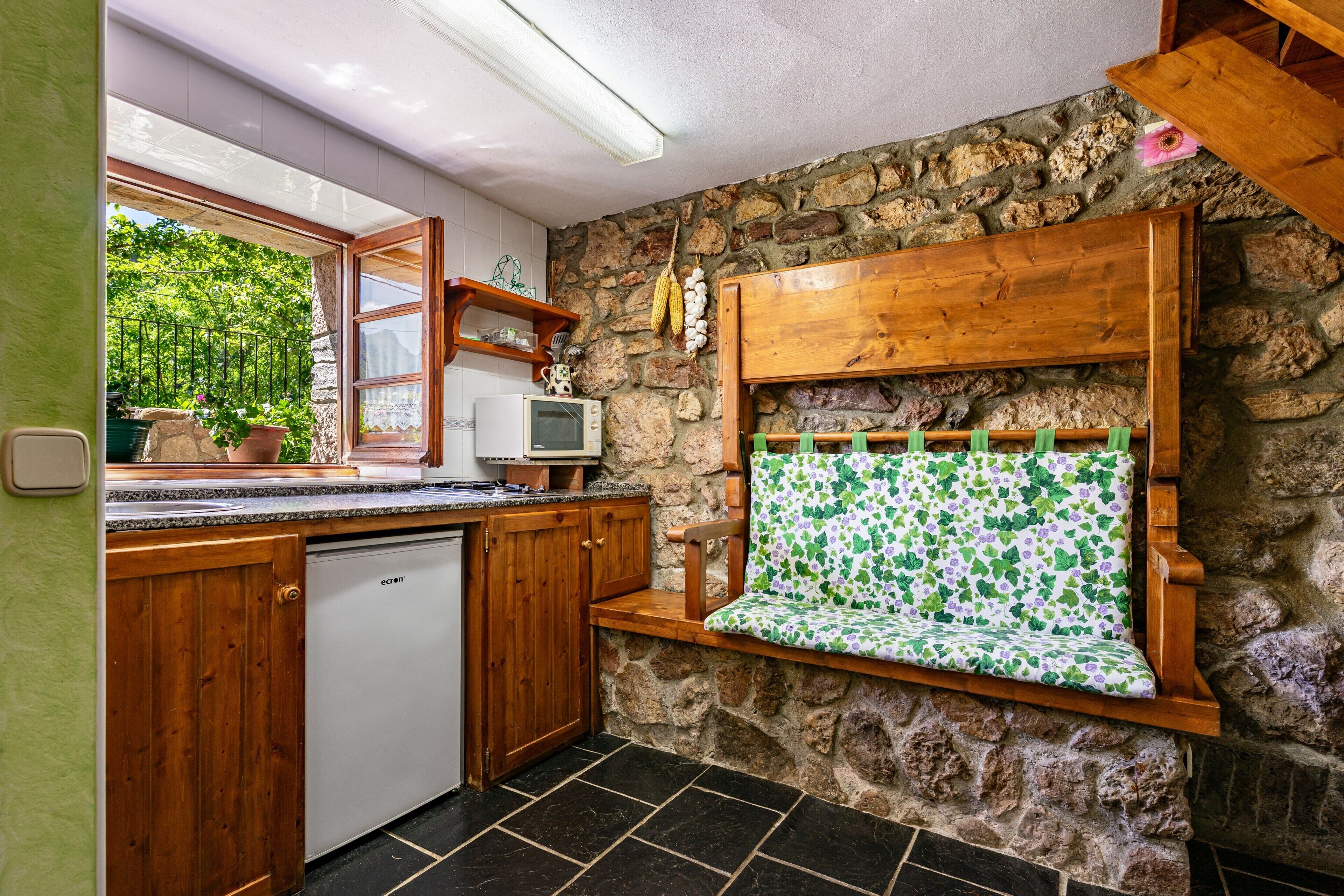 Una cocina con pared de piedra, armarios de madera, un banco empotrado con cojín floral, un televisor pequeño y una ventana con vistas a la vegetación.