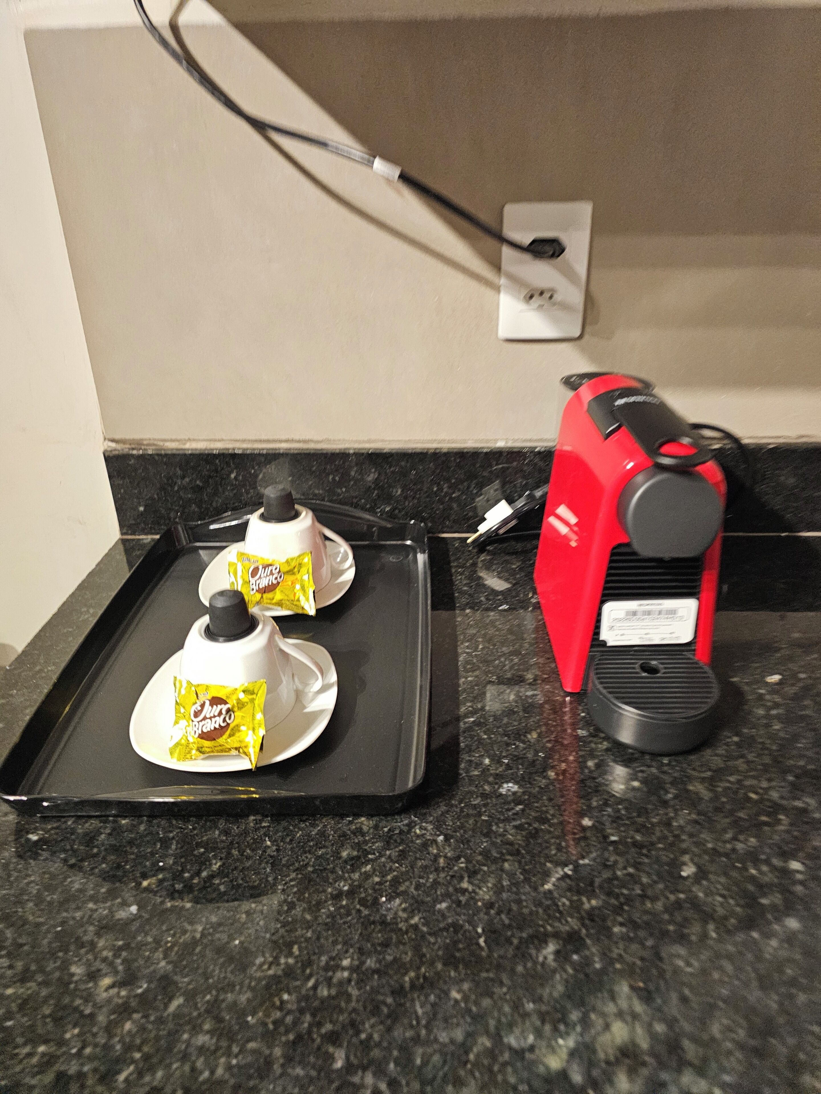 Una encimera de cocina con una cafetera, una bandeja con dos sobres de café y un enchufe en la pared.