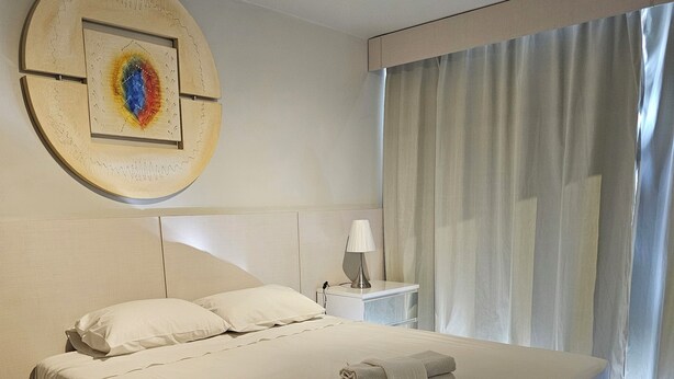 Ein ordentlich bezogenes Bett mit weißen Laken, ein Nachttisch mit Lampe und ein an der Wand befestigtes Kunstwerk.