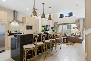 Eine moderne Küche mit einer zentralen Kochinsel, Barhocker, einem Essbereich mit einer Couch und einem Wohnzimmer mit Fernseher und Pflanzen.