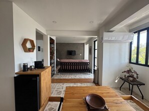 A modern room with a wooden dining table, a bed with a pink bedspread, a TV mounted on the wall, and a window with a view of trees. - Apartment with AC in Colonia Reforma (pet friendly) 30 minutes from downtown (Oax.)