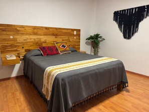 A bedroom with a wooden headboard, a grey bedspread, two patterned pillows, a bedside plant, and a wall hanging. - Apartment with AC in Colonia Reforma (pet friendly) 30 minutes from downtown (Oax.)