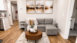 A modern living room with a grey sectional sofa, a round wooden coffee table, and abstract wall art. - Cozy & Spacious 2BR Private Suite Near ATL Airport (Atlanta)