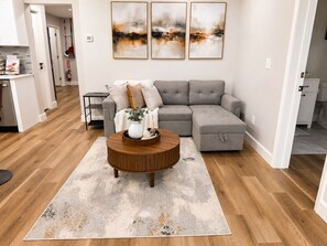 Ein modernes Wohnzimmer mit einem grauen Ecksofa, einem runden Couchtisch aus Holz und abstrakter Wandkunst.