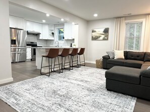 A modern kitchen with stainless steel appliances, a central island with bar stools, a dark grey sofa, and a patterned rug. - Idyllic Cottage In Campobello by Tryon International (Campobello)